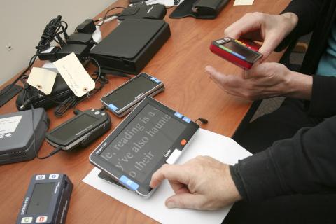 A tabletop covered with various Assistive Technology devices, including Braille displays and notetakers.