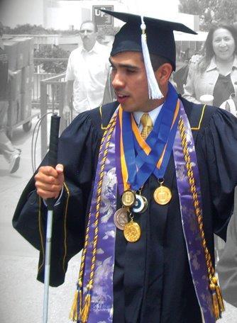 A young man with a white cane wearing a cap and gown at graduation ceremony.