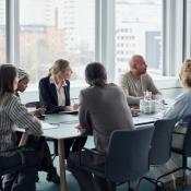 People sitting around a conference room table. They are listening and taking notes as a woman speaks.