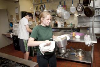 A teenager carries a large pot in a commercial kitchen. Another teen is preparing food on the counter behind them.