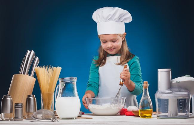 A young girl wearing an apron and chef’s hat uses a whisk to stir ingedients in a bowl.