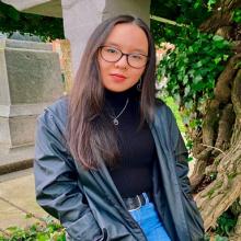 May is Vietnamese, she has long black hair and she has black glasses. She is wearing a black turtleneck with jeans and a black jacket. She is wearing silver earrings, a silver necklace, and a black belt with a silver buckle. There is a stone structure on her left and a tree on her right.