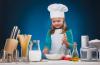A young girl wearing an apron and chef’s hat uses a whisk to stir ingedients in a bowl.